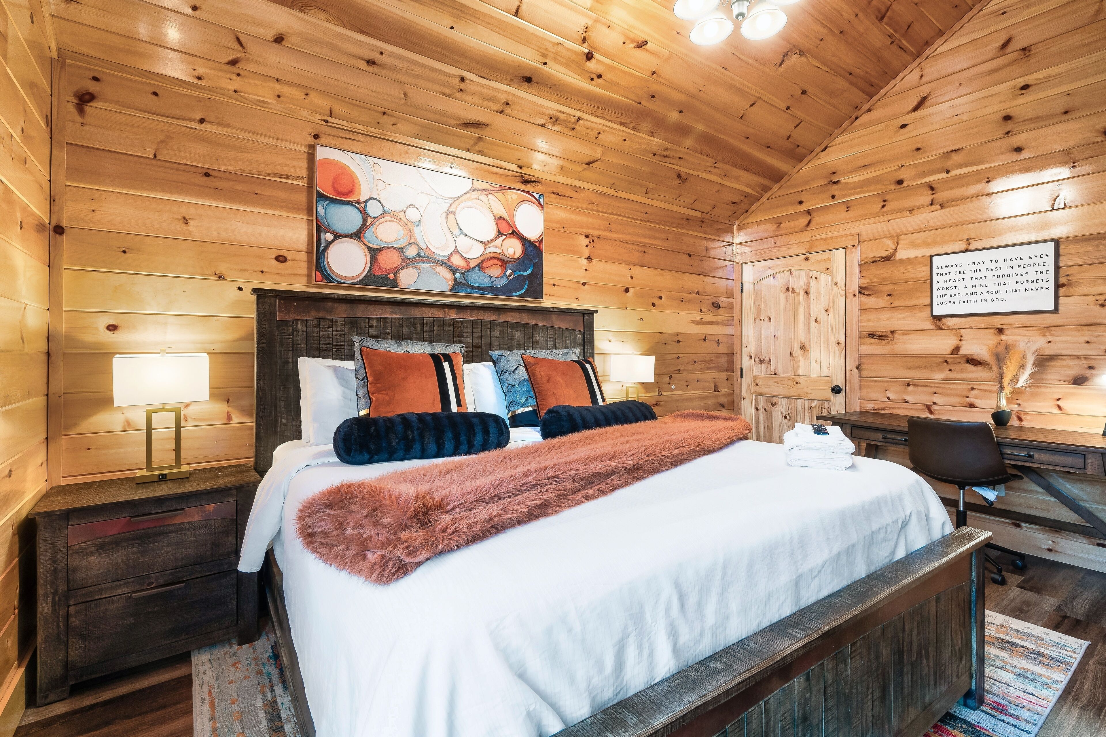 A wooden cabin bedroom with a large bed, a headboard with a painting, a desk with a chair, and a wall-mounted lamp.