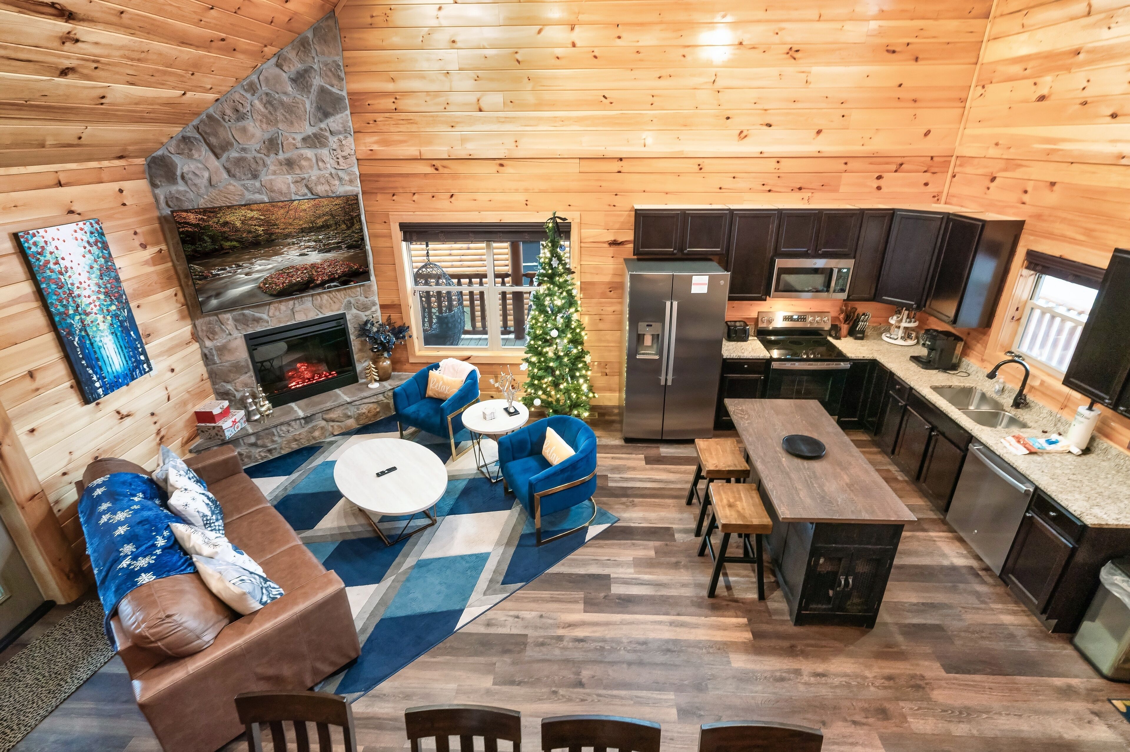 Interior acogedor de una cabaña con chimenea de piedra, sofás azules, comedor y cocina con armarios oscuros y electrodomésticos de acero inoxidable.