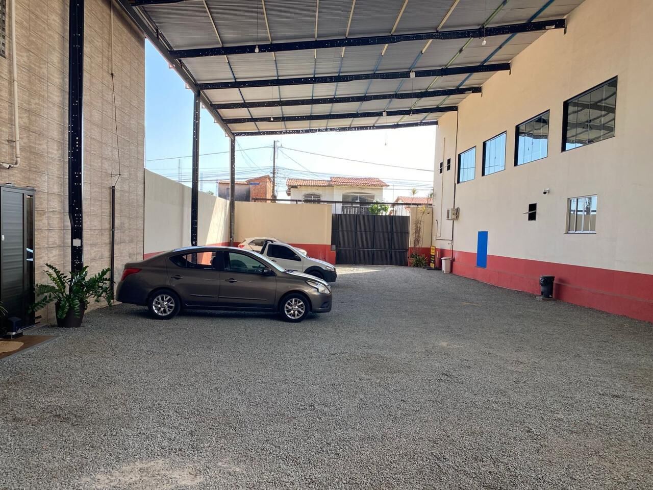 Un área de estacionamiento techada con dos coches aparcados, un edificio de fachada roja y blanca, y cielo despejado.