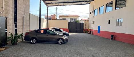 Un área de estacionamiento techada con dos coches aparcados, un edificio de fachada roja y blanca, y cielo despejado.