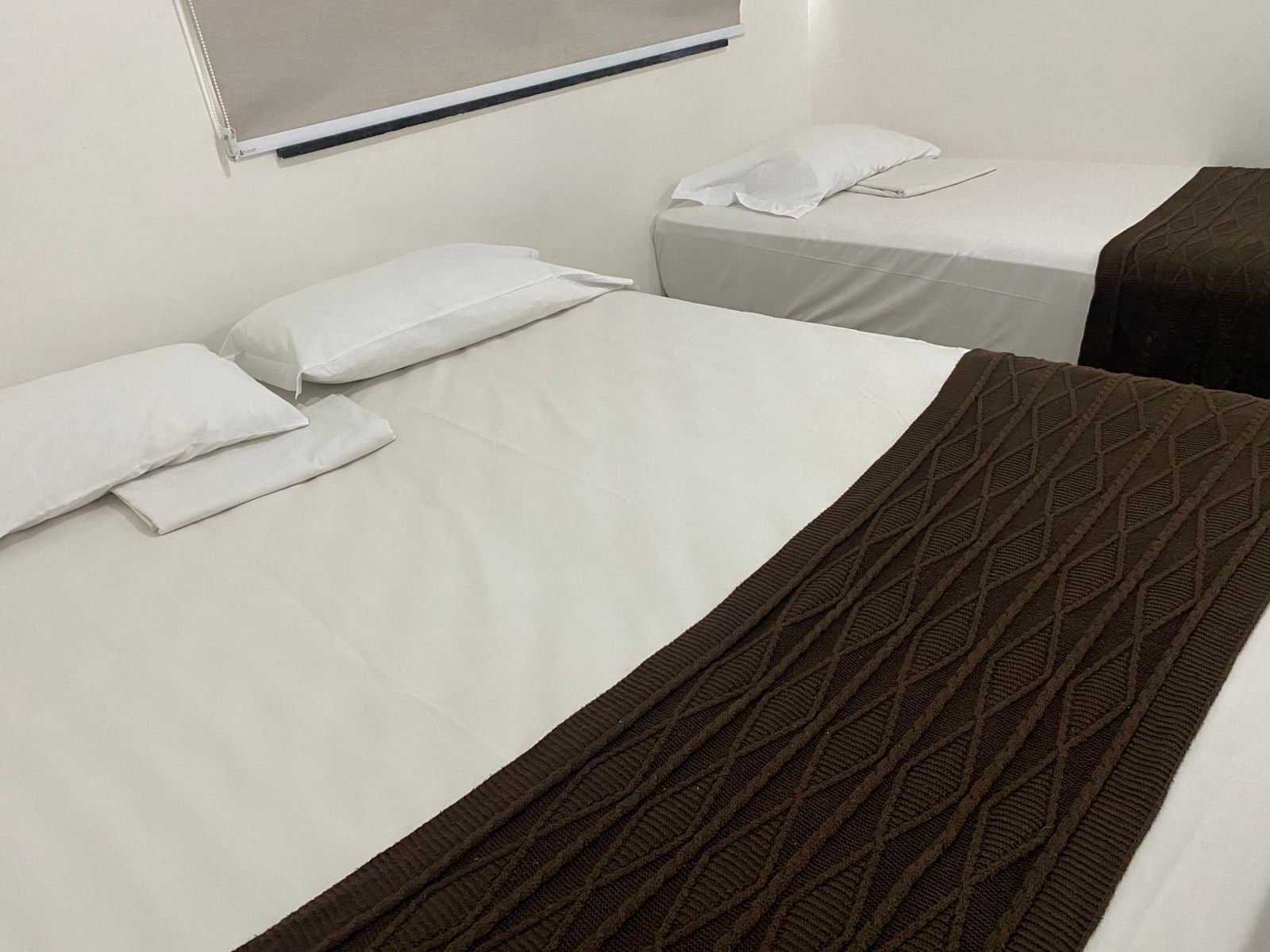 Habitación de hotel con dos camas, cada una con sábanas y almohadas blancas. Una cama tiene una manta marrón.