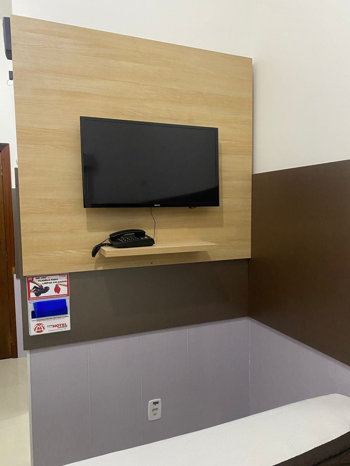 A hotel room with a mounted flat-screen TV, a telephone, and a small shelf.