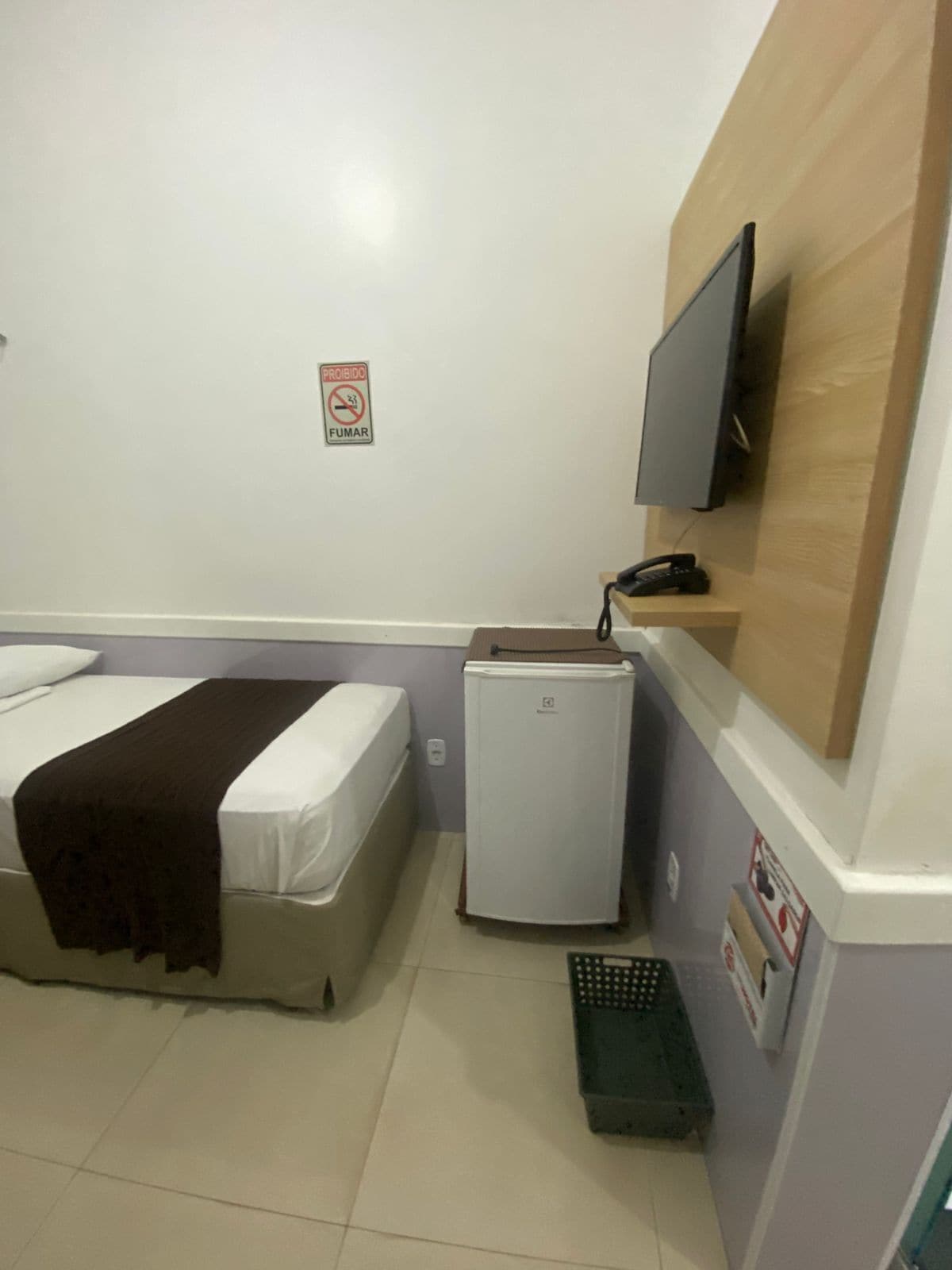 A hotel room with a bed, a small refrigerator, a television mounted on the wall, and a no-smoking sign.