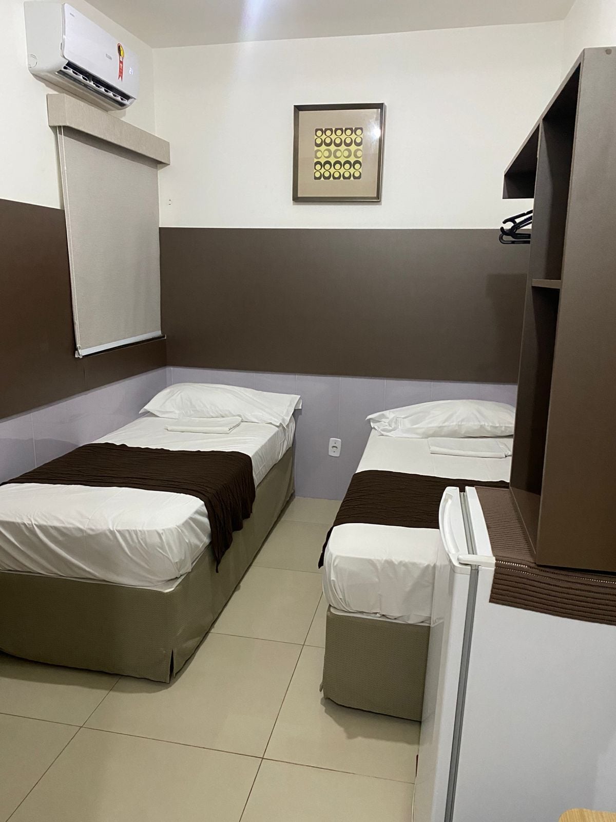 A small, compact room with two single beds, a white air conditioning unit, a framed picture on the wall, and a small kitchenette area.