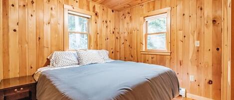 Une chambre à coucher aux murs lambrissés, avec un lit, deux fenêtres et un ventilateur de plafond.