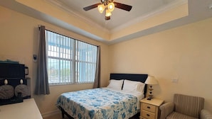 Un dormitorio con cama, una silla, una mesita de noche, un escritorio, una ventana con persianas y un ventilador de techo, con vistas al exterior.