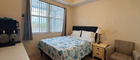Un dormitorio con cama, una silla, una mesita de noche, un escritorio, una ventana con persianas y un ventilador de techo, con vistas al exterior.