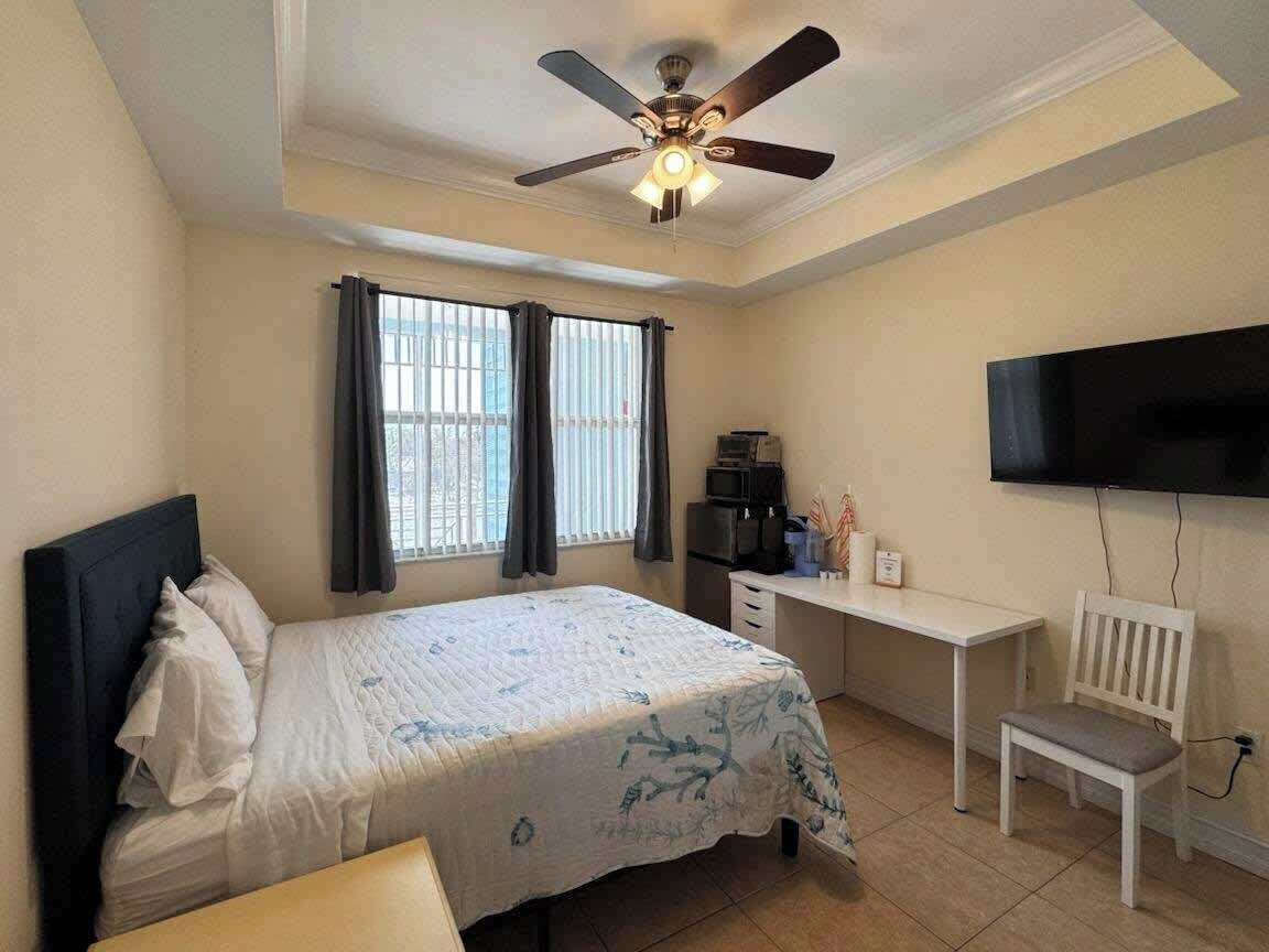 A bedroom with a bed, a desk, a chair, a ceiling fan, and a TV.