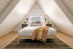 Dormitorio tipo loft con techo inclinado, cama con estructura de madera, dos mesitas de noche y vistas a zonas verdes.