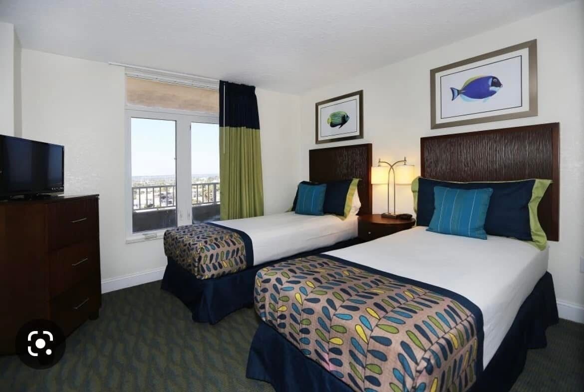 A hotel room with two beds, a TV, a window with curtains, and artwork on the walls.
