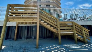 En trækonstruktion med flere niveauer og en trappe, der fører op til en balkon, mod en klar blå himmel.