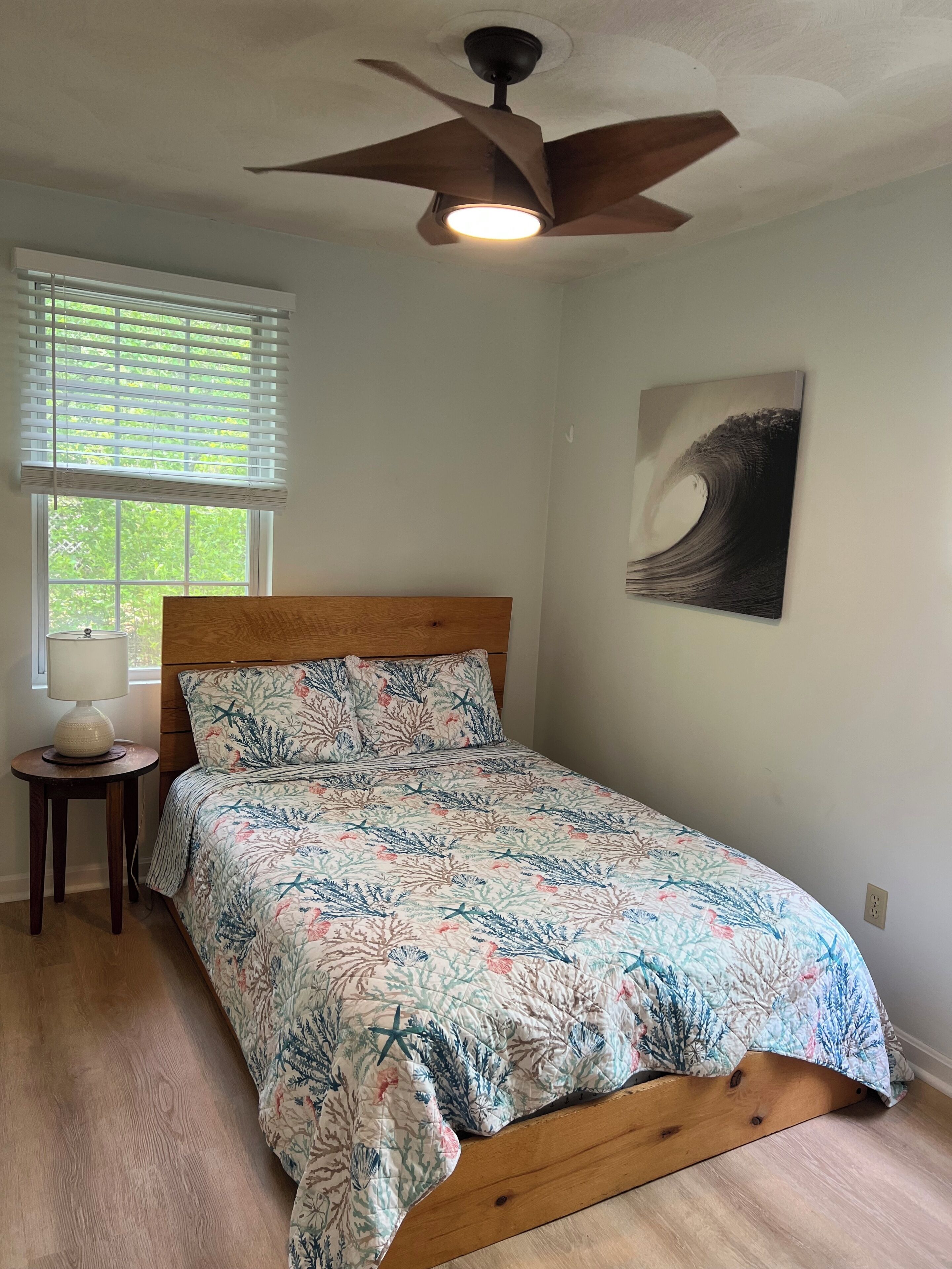 Quarto com cama, ventoinha no teto, janela com persianas, luminária em mesa de apoio e pintura na parede.