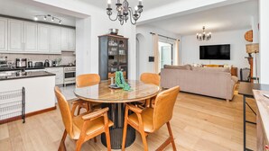 Une cuisine moderne avec des meubles blancs, une table à manger avec des chaises, un lustre et un coin salon avec un canapé et un téléviseur.