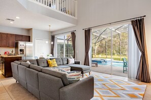 A spacious living area with a grey sectional sofa, a kitchen with dark wood cabinets, and a large sliding glass door leading to a pool. - 6BR Disney Villa, Heated Pool and Game Room (Kissimmee)