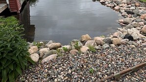 En stuga vid sjön med en brygga som sträcker sig ut i det stilla vattnet, omgiven av en klippig strand och grönskande vegetation.