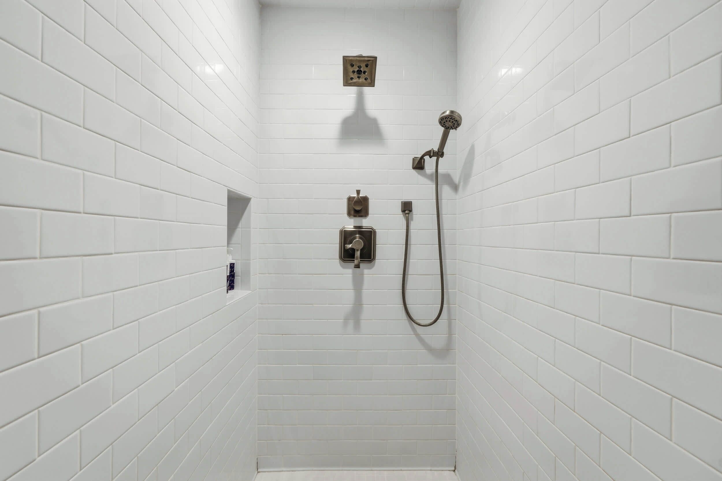 Un espace douche avec des murs recouverts de carreaux blancs, une douchette à main et un robinet fixé au mur.