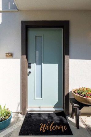 A front door with a 'Welcome' mat, a potted plant, and a small table with a flower pot.