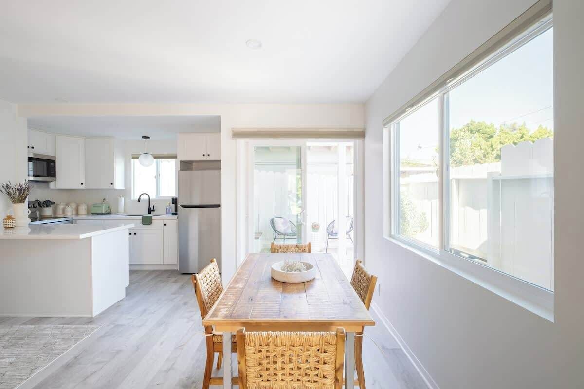Una cocina moderna con armarios blancos, un comedor con mesa de madera y sillas de mimbre, y ventanas amplias que ofrecen luz natural.