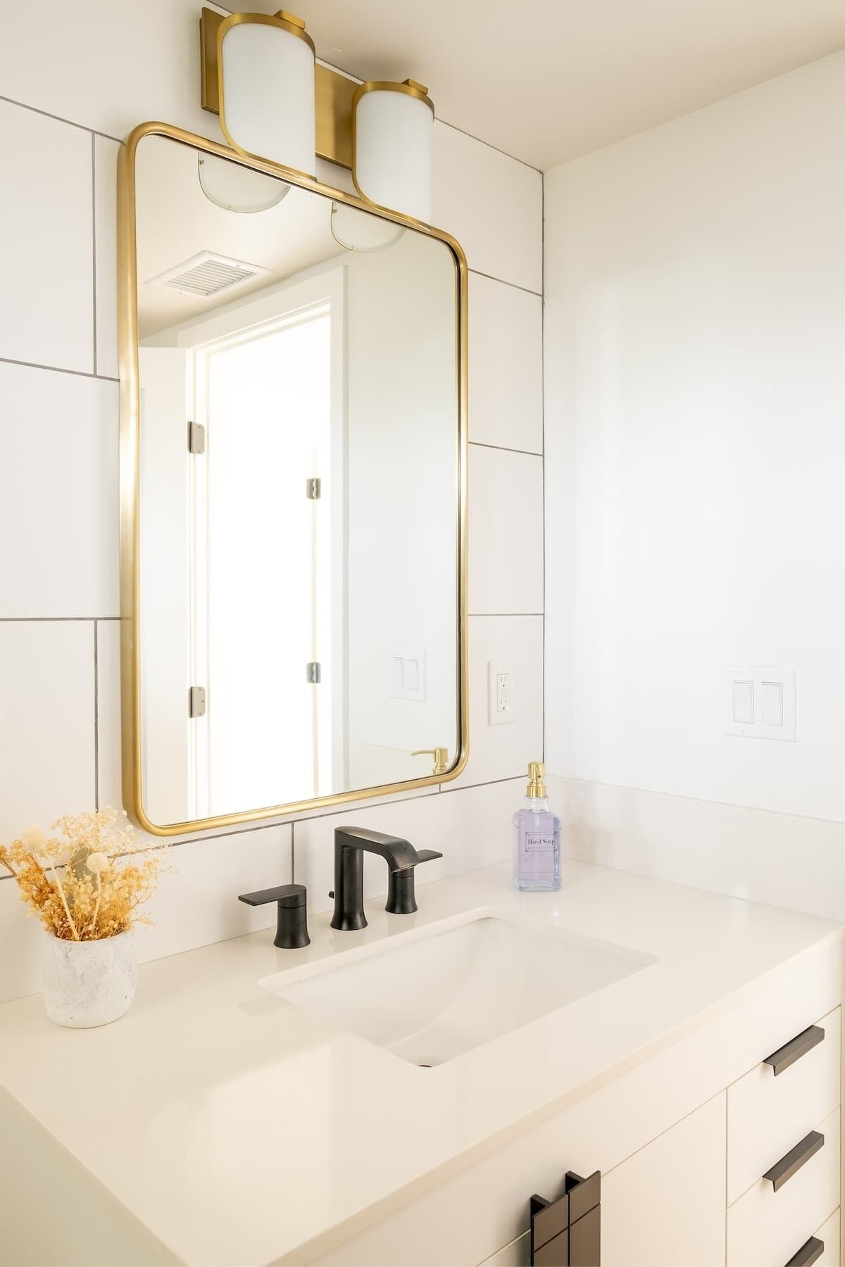 A modern bathroom with a white vanity, a gold-framed mirror, a black faucet, and a small vase with dried flowers.