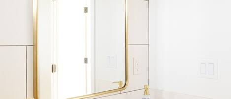 A modern bathroom with a white vanity, a gold-framed mirror, a black faucet, and a small vase with dried flowers.