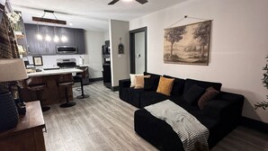 Una sala de estar moderna con un sofá negro, piso de madera, un ventilador de techo y una cocina al fondo.
