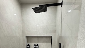 Un área de ducha moderna con estante de vidrio para artículos de aseo, cabezal de ducha manual y desagüe en el suelo.