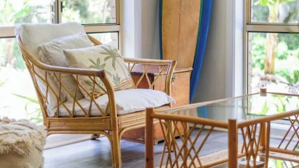 Een rieten stoel met kussens, een houten surfboard tegen een muur geplaatst, en een glazen tafel in een kamer met grote ramen.