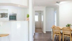 Een moderne keuken met witte kastjes, een inbouwoven en een zithoekje met rieten stoelen en een houten tafel.