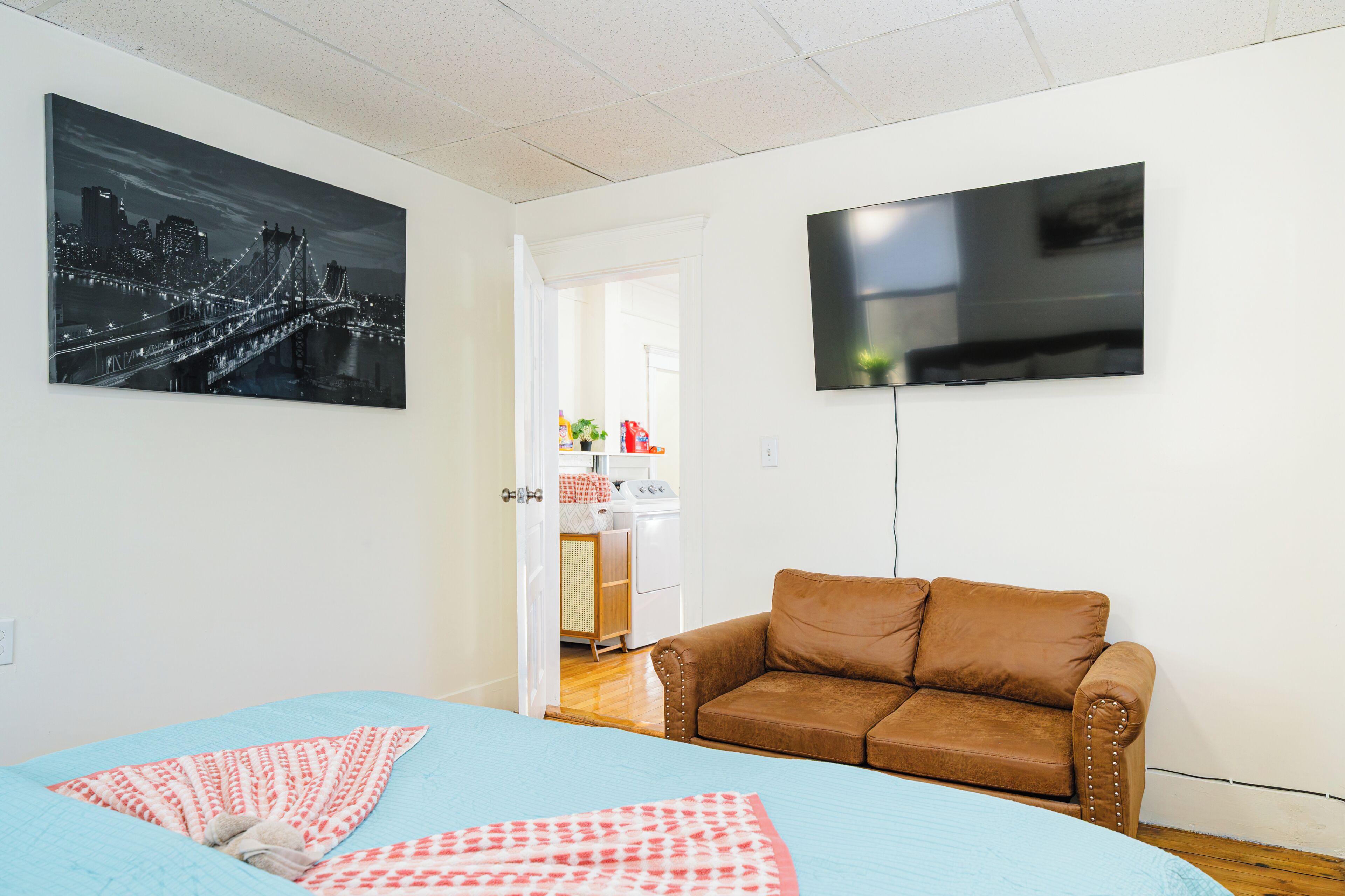 Um quarto com uma cama, um sofá de couro marrom, uma televisão de tela plana e uma pintura com vista para a cidade na parede.