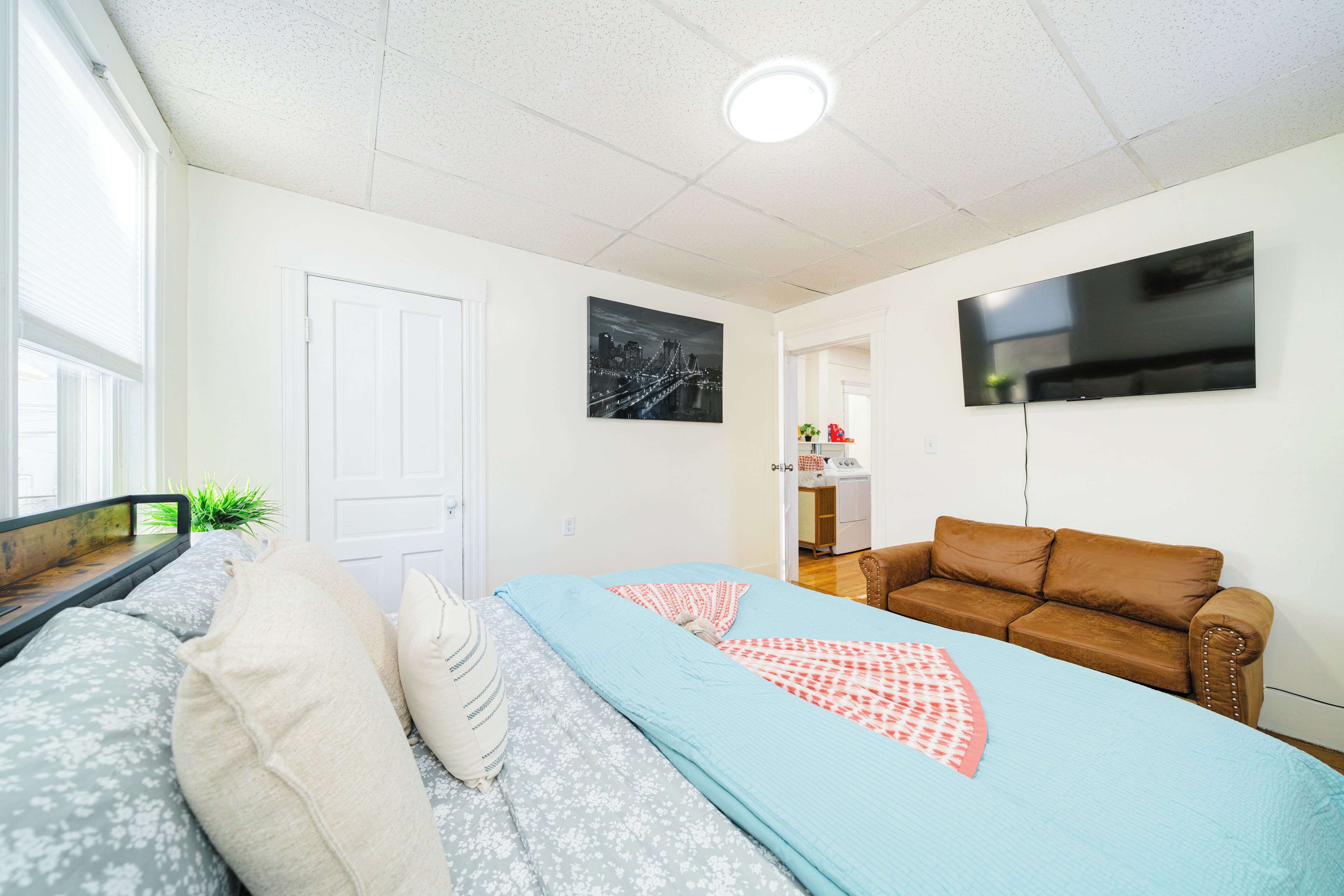 A bedroom with a bed, a brown leather sofa, a flat-screen TV mounted on the wall, and a door.