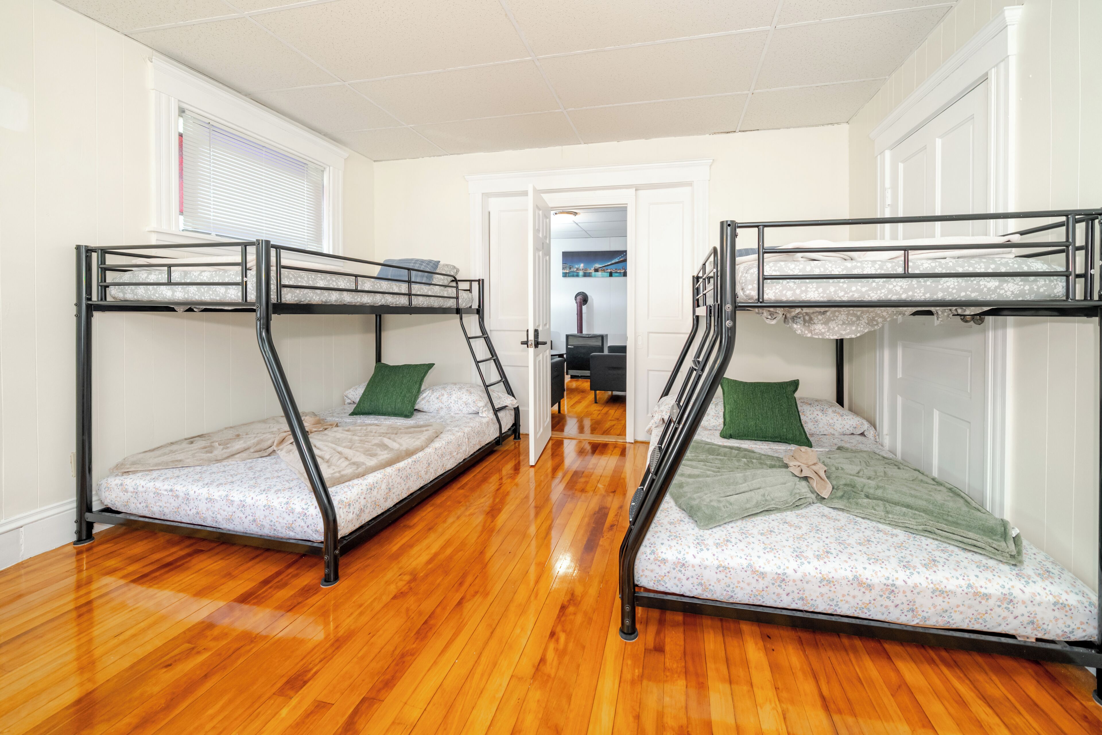 A room with two bunk beds, wooden flooring, a door leading to another room, and a window with blinds.