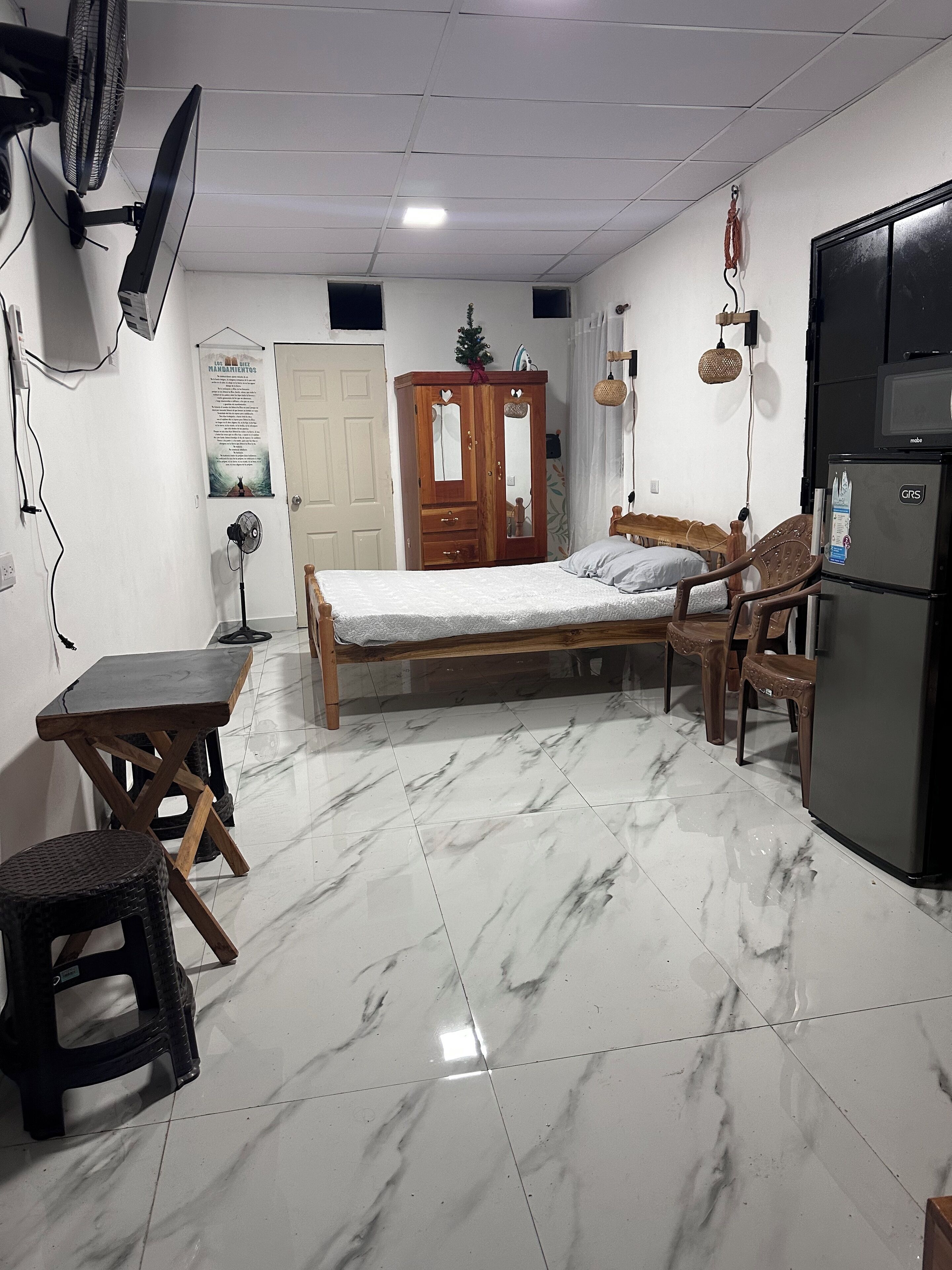 A room with a bed, a wooden wardrobe, a small table, a fan, a refrigerator, and a tiled floor.