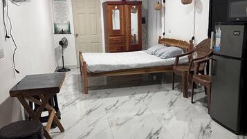 A room with a bed, a wooden wardrobe, a small table, a fan, a refrigerator, and a tiled floor.