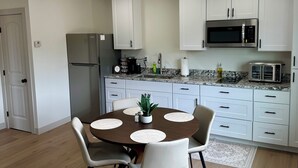 Une cuisine moderne avec des armoires blanches, un four à micro-ondes encastré et un réfrigérateur. On y trouve une table à manger avec quatre chaises, et une plante en pot posée dessus.