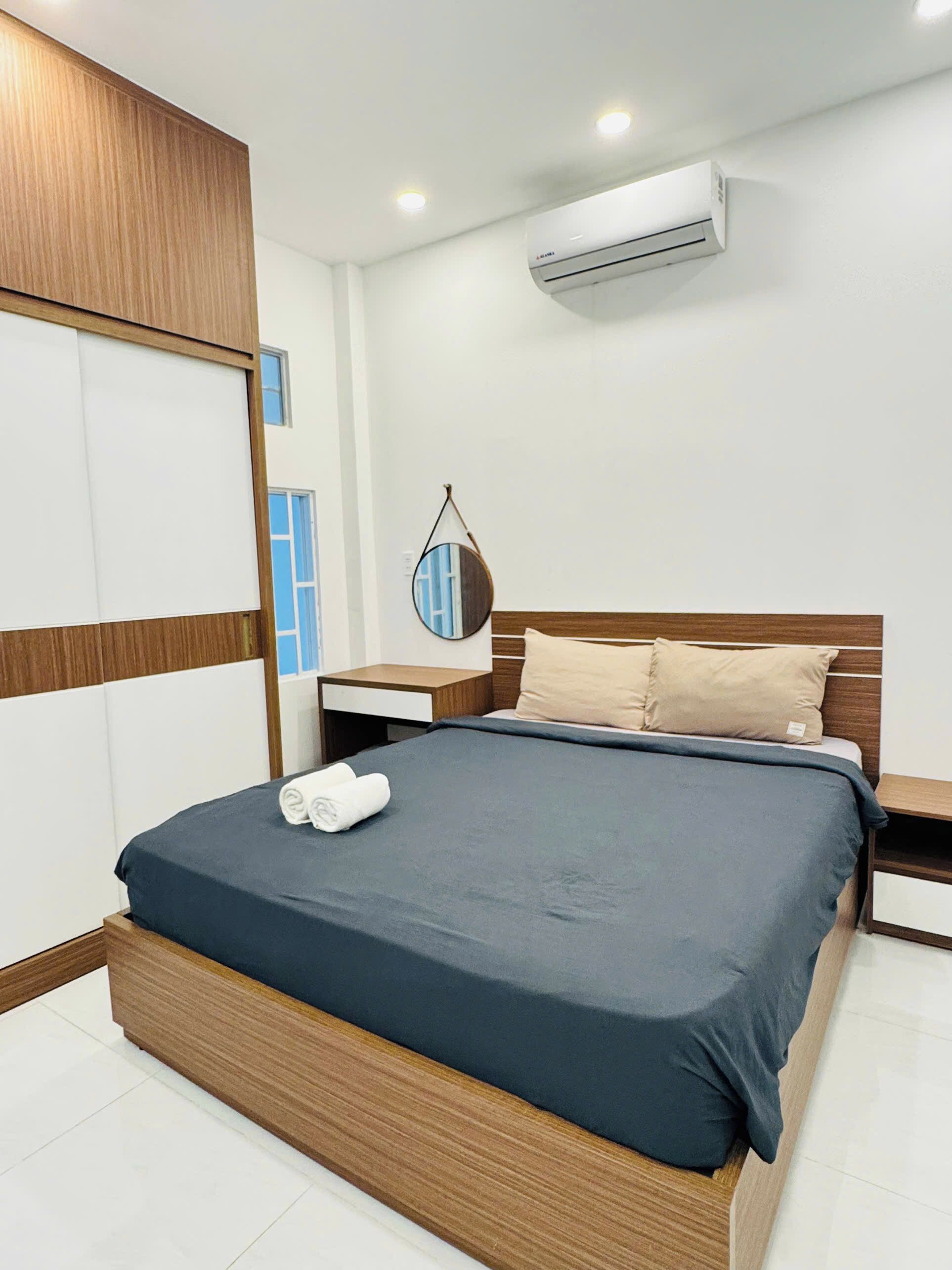 A modern hotel room with a wooden headboard, a large bed with dark bedding, a bedside table, a nightstand, a wall-mounted air conditioner, and a mirror with a circular frame.