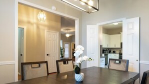 A dining room with a dark wooden table and chairs, a white vase with flowers, and a kitchen visible through an open door. - Suburban Retreat Central to Dallas, Plano & Frisco (Dallas)