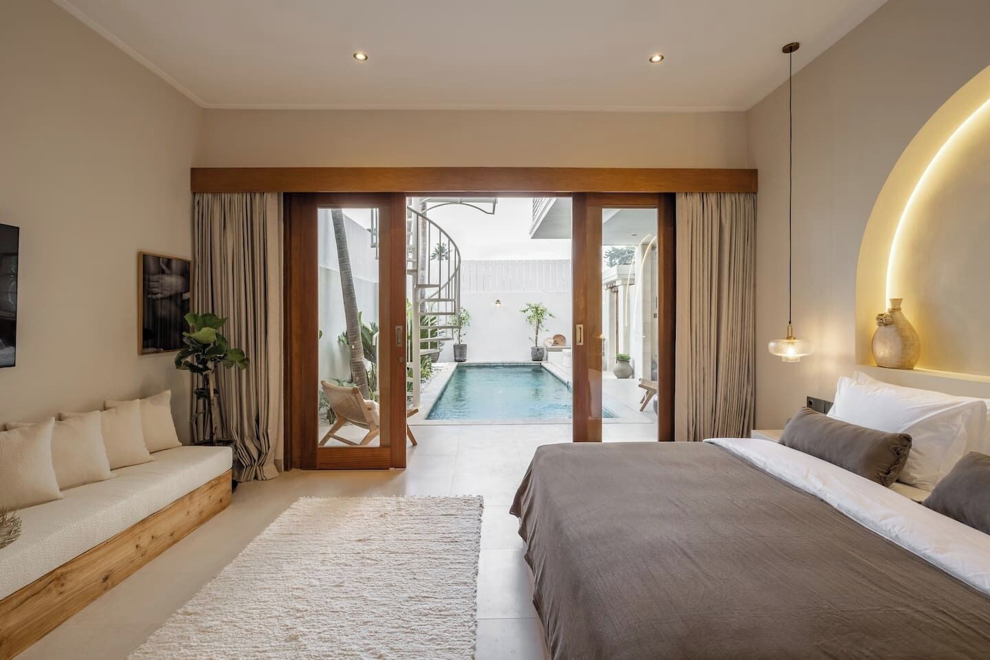 A modern bedroom with a large bed, a sofa, a pool view, and a staircase leading to an outdoor area.