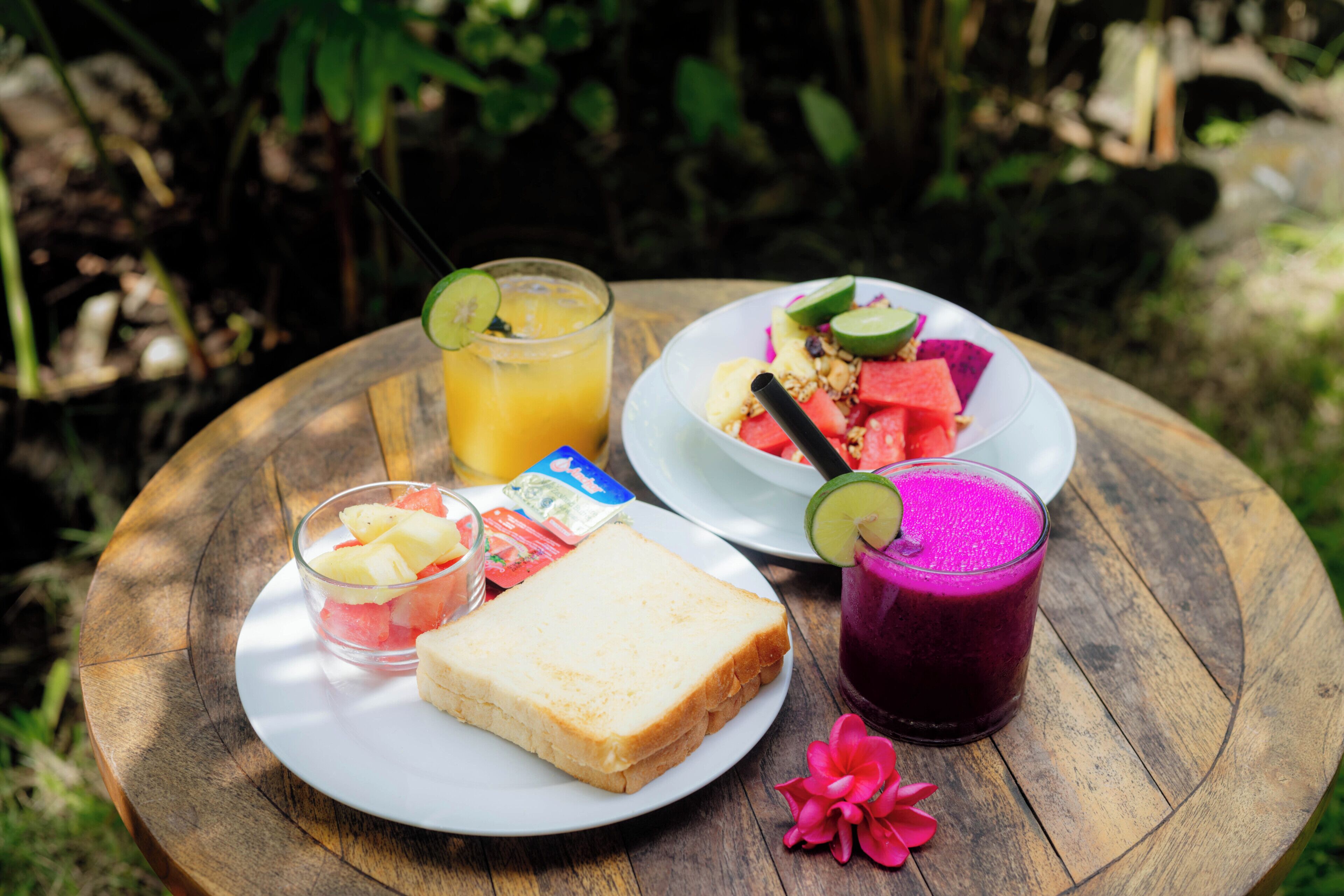 A wooden table with a plate of toast, a glass of orange juice, a bowl of fruit, a plate with watermelon and granola, and a glass of purple smoothie.