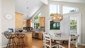 A kitchen with wooden cabinets, a dining table with chairs, a clock, and a window with blinds. - Harbor Haven-Ocean Views, Steps to Beach + Dining (Santa Cruz)