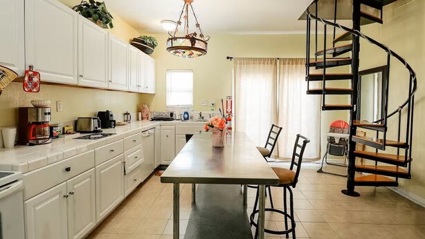 Une cuisine avec des meubles blancs, une table à manger, des chaises, un lustre et un escalier en colimaçon.