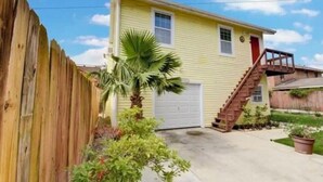 Une maison jaune d’un seul étage, avec une clôture en bois, un palmier et un escalier menant à un garage.