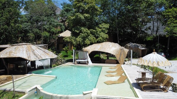 Un área de piscina con sillones de descanso y sombrillas, rodeada de árboles y estructuras de madera.