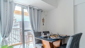 A dining area with a table set for two, a balcony with wicker chairs, and a view of buildings outside. - Apartment in Santos, 2 bedrooms, brand new, decorated, pet friendly - Aloha 181. (Santos)