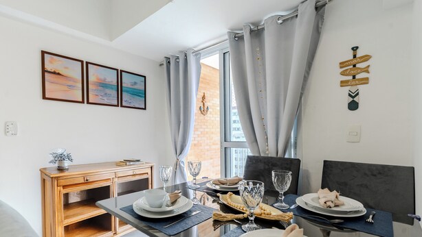 A dining area with a table set for two, a wooden sideboard, and a view of the outside through a window with curtains. - Apartment in Santos, 2 bedrooms, brand new, decorated, pet friendly - Aloha 181. (Santos)