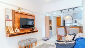 A modern living room with a flat-screen TV, grey sofa, and ottoman. A kitchen with a refrigerator and cabinets is visible in the background. - Apartment in Santos, 2 bedrooms, brand new, decorated, pet friendly - Aloha 181. (Santos)