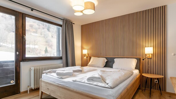 A modern bedroom with a wooden bed, a large window, and a wall-mounted lamp.