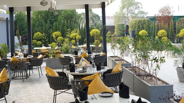 An outdoor dining area with round tables and chairs, surrounded by greenery and a covered walkway. - JBM RESORTS & WATER PARK (Abohar)