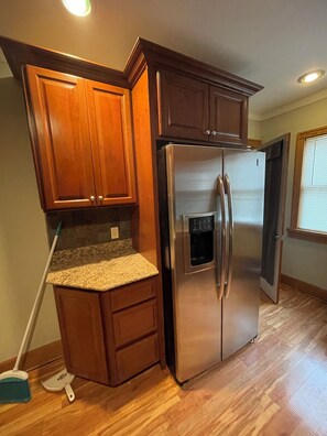 A kitchen with wooden cabinets, a stainless steel refrigerator, granite countertops, and a broom leaning against the cabinets. - Mid-Term Rental - Over 30 Days - Saint Paul near colleges (Saint Paul)