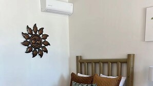 A neatly made bed with a brown quilt and pillows, a wooden headboard, a wall-mounted air conditioner, and a decorative sun-like wall piece. - Costa del Sol Hotel & Sportfishing (Bahía de los Ángeles)