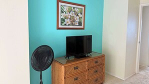 Een kamer met blauwgroene wanden, een houten dressoir met lades, een zwarte televisie erboven en een staande ventilator.